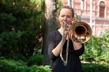 Girl learning to play trombone. Portrait of a girl on a background of a city park park. Portrait. Close-up.の写真素材