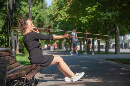 Girl learning to play trombone. Girl plays sitting on a park bench. Portrait.の写真素材