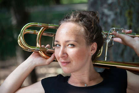Girl learning to play trombone. Portrait of a girl on a background of a city park park. Portrait. Close-up.の写真素材