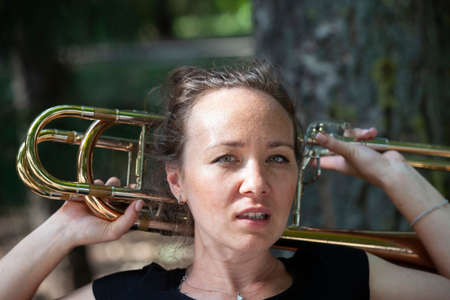 Girl learning to play trombone. Portrait of a girl on a background of a city park park. Portrait. Close-up.の写真素材