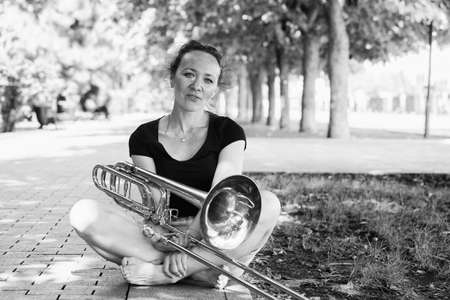 Girl learning to play trombone. Girl plays sitting on the plates of a park alley. Portrait.の写真素材