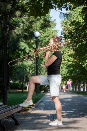 Girl learning to play trombone. Girl plays standing on the alley of a city park. Portrait.の写真素材