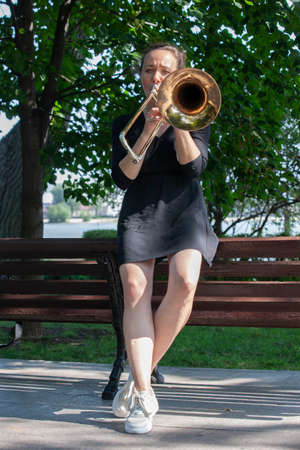 Girl learning to play trombone. Girl plays leaning on a park bench. Portrait.の写真素材