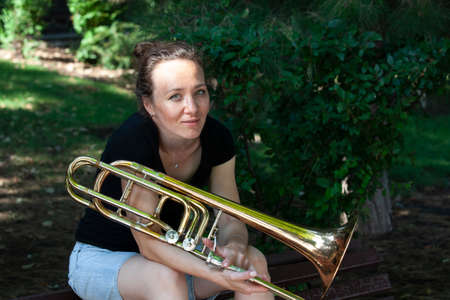 Girl learning to play trombone. Portrait of a girl on a background of a city park park. Portrait. Close-up.の写真素材