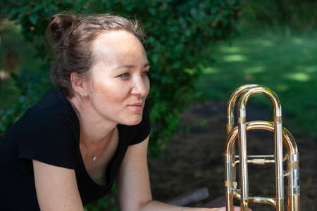 Girl learning to play trombone. Portrait of a girl on a background of a city park park. Portrait. Close-up.の写真素材