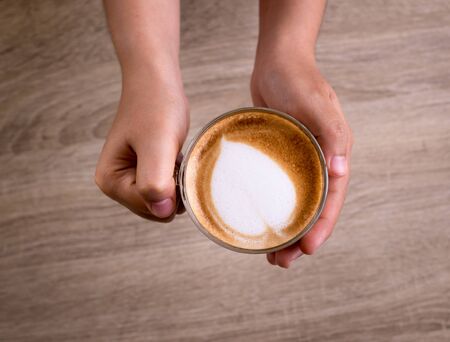 Female hands holding a cup of coffee with heart make up on face of milk cream on wooden table, top viewの写真素材
