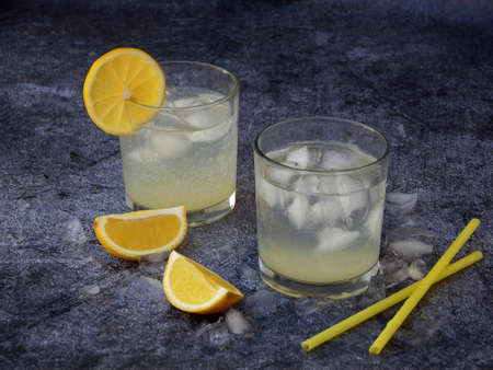 glasses of cold homemade lemonade with lemon slices, ice cubes and straws on dark background. Copy...