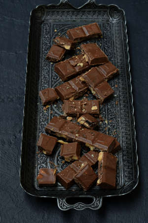 Milk chocolate with nuts and dried fruits on a metal tray on a dark concrete background.