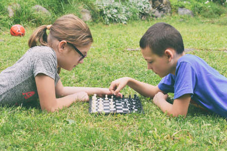 Girl and boy playing chess outdoors. Children lie on green grass in summer park. Happy childhood.