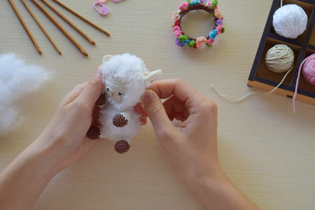 Making fanny sheep with wreath of flowers. Crochet toy ewe for child. On table threads, needles,...