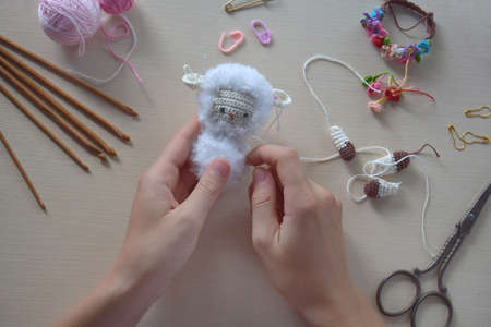 Making fanny sheep with wreath of flowers. Crochet toy ewe for child. On table threads, needles,...