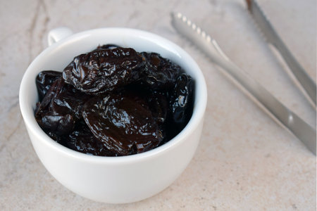Dried Prunes in White Bowl with Fork on Neutral Background. Healthy Snack for Vegan Diet, Rich in Fiber and Nutrients, Ideal for Culinary Recipes, Desserts, and Weight Managementの写真素材