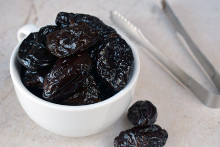 Dried Prunes in White Bowl with Fork on Neutral Background. Healthy Organic Snack for Vegan Diet, Rich in Fiber and Nutrients, Ideal for Culinary Recipes, Desserts, and Weight Managementの写真素材