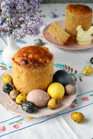 Delicious Easter Kulich with Colorful Eggs and Blossoming Lilac Flowers. Traditional Holiday Pastry Presented on Tablecloth with Spring Elements and Natural Light for Festive Celebrationの写真素材
