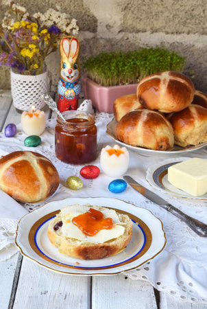 Festive Easter Breakfast with Hot Cross Buns, Butter, Jam, Chocolate Bunny, Painted Eggs, Fresh Flowers, Rustic White Wooden Table Setting, Holiday Pastry, Natural Spring Decorationの写真素材