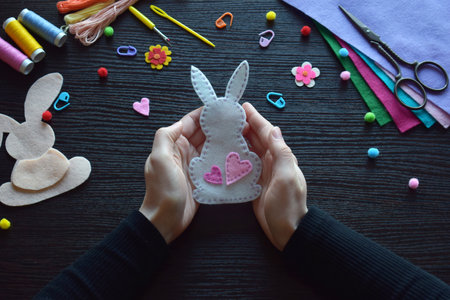 Displaying finished felt bunny ornament, Easter handmade decoration, DIY craft project, festive accessory, colorful workspace materials, spring holiday preparation, threads, buttons, creative processの写真素材