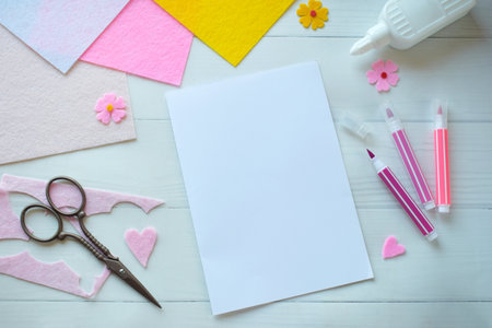Creative flat lay composition of DIY greeting card crafting materials, including colorful felt flowers, scissors, glue, and blank paper, arranged on a white wooden background, showing the process of making a personalized handmade card for special occasionsの写真素材