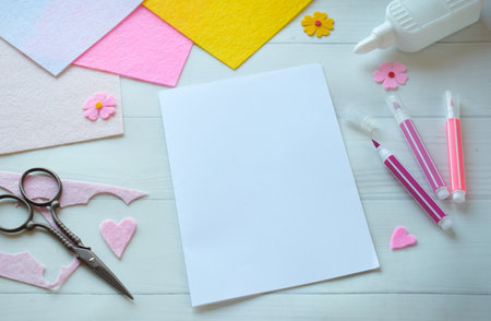 Creative flat lay composition of DIY greeting card crafting materials, including colorful felt flowers, scissors, glue, and blank paper, arranged on a white wooden background, showing the process of making a personalized handmade card for special occasionsの写真素材