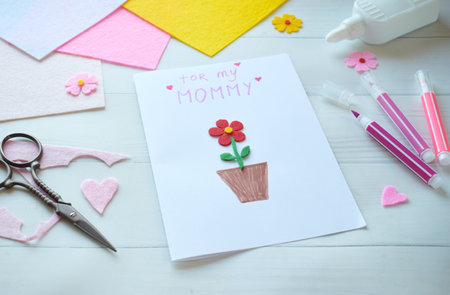 Handmade Mothers Day Greeting Card Creation. DIY Craft Project with Felt Flowers, Colorful Paper, and Scissors on White Wooden Background. Creative Holiday Gift Idea for Mom, Festive Cardmaking Process with Crafting Suppliesの写真素材