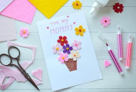 Handmade Mothers Day Greeting Card Creation. DIY Craft Project with Felt Flowers, Colorful Paper, and Scissors on White Wooden Background. Creative Holiday Gift Idea for Mom, Festive Cardmaking Process with Crafting Suppliesの写真素材