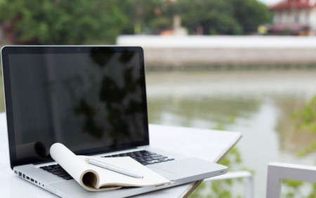 Blank notepad and pen over laptop next to the river on the tableの写真素材