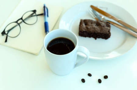 Coffee with blank paper note and glasses and chocolate brownie on white tableの写真素材