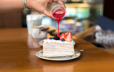 Hand pouring strawberry sauce over crape cake with fresh fruit on topの写真素材