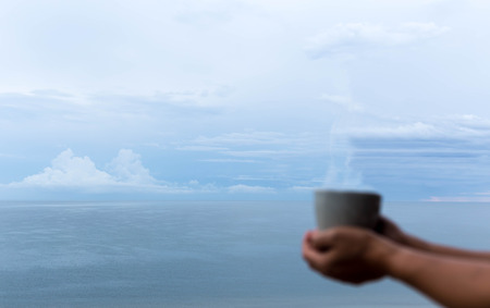Blurred hand holding coffee mug with soft ocean view as backgroundの写真素材