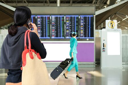 Woman using mobile phone looking at flight timetable with flight attendants walking in airport as backgroundの写真素材