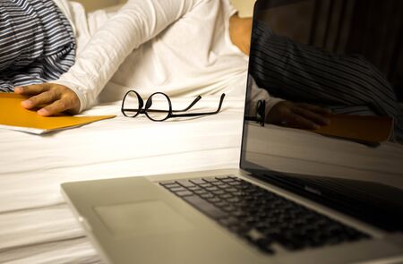 Focus on glasses businessman working on laptop fall sleep on the bed with hand holding bookの写真素材