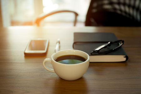 Cup of coffee and note pad with pen and glasses on the wooden desk with sunlightの写真素材