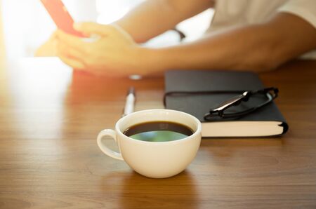 Focus on coffee cup man looking on cell phone in background with notebook and glasses on the tableの写真素材