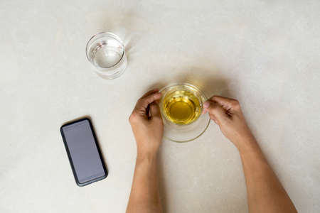 Top view hand holding cup of tea with cell phone, glass of water on tiled floor backgroundの写真素材