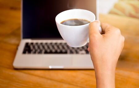 Hand holding a coffee cup with laptop sitting on the tableの写真素材