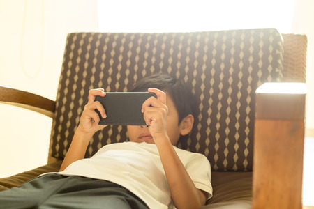 Asian boy using cell phone and laying down on chair with light flare in backgroundの写真素材
