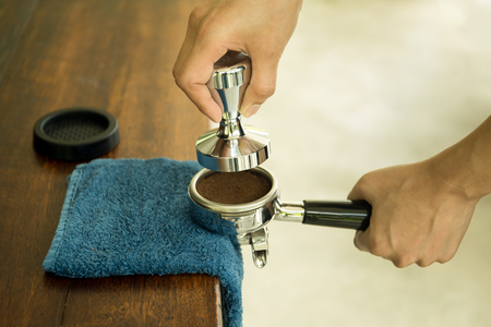 Barista pressing ground coffee into portafilter using tamper with blue cloth on the tableの写真素材