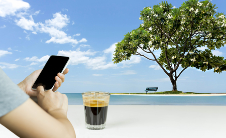 Hand using cell phone and coffee with beautiful frangipani tree with chair on the beach on vacationの写真素材