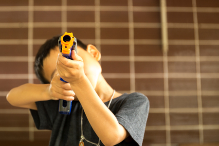 Little boy playing with a toy gun against orange brick wall backgroundの写真素材