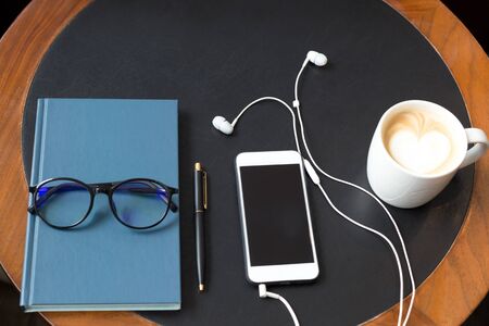 Cell phone with headphones and glasses on notebook and pen lying with coffee on the tabelの写真素材