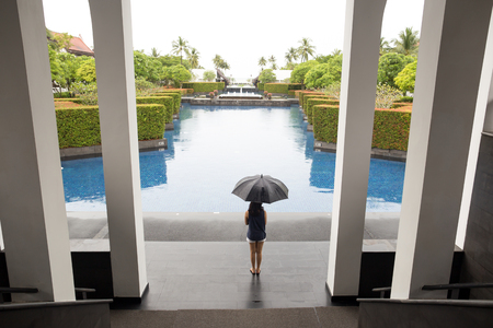 Woman with umbrella standing by swimming pool watching the rain on vacationの写真素材
