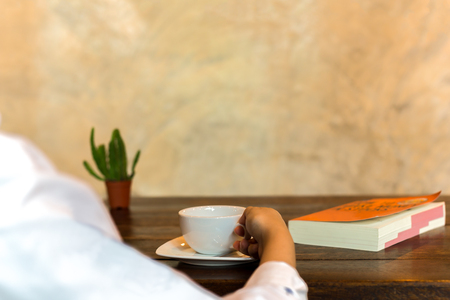 Hand holding coffee cup with book on wooden table and small plantの写真素材
