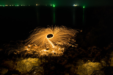 Man showers of glowing sparks from spinning steel wool on rocky beach at nightの写真素材
