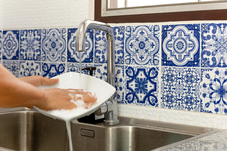 Hands washing white plate in kitchen sink with running water with blue tileの写真素材