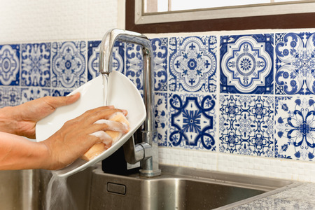 Hands washing white plate in kitchen sink with running water with blue tileの写真素材