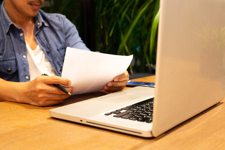 Man hand holding pen reading paper work with laptop on the tableの写真素材