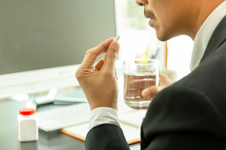 Businessman taking pills at work desk in office with glass of water for health careの写真素材