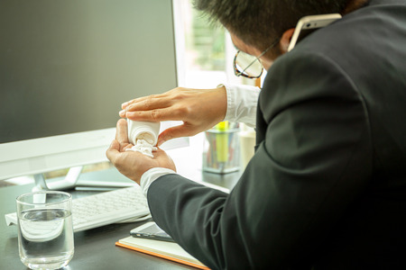 Businessman busy talking on cell phone and taking pills at work desk in office with glass waterの写真素材