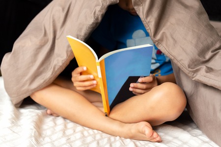 Young boy reading a book under the blanket in bed.