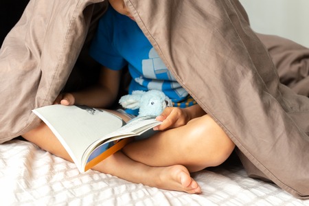 Young boy reading a book under the blanket in bed.の写真素材