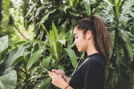Mixed race teenage woman with earphones listening to the music, against green trees tropical background.の写真素材